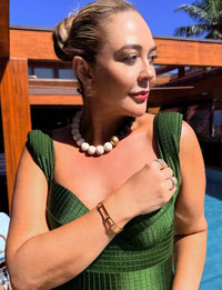 Mulher elegante com maiô feminino modelador verde, acessórios de joias e penteado sofisticado em ambiente ao ar livre com piscina e plantas ao fundo.