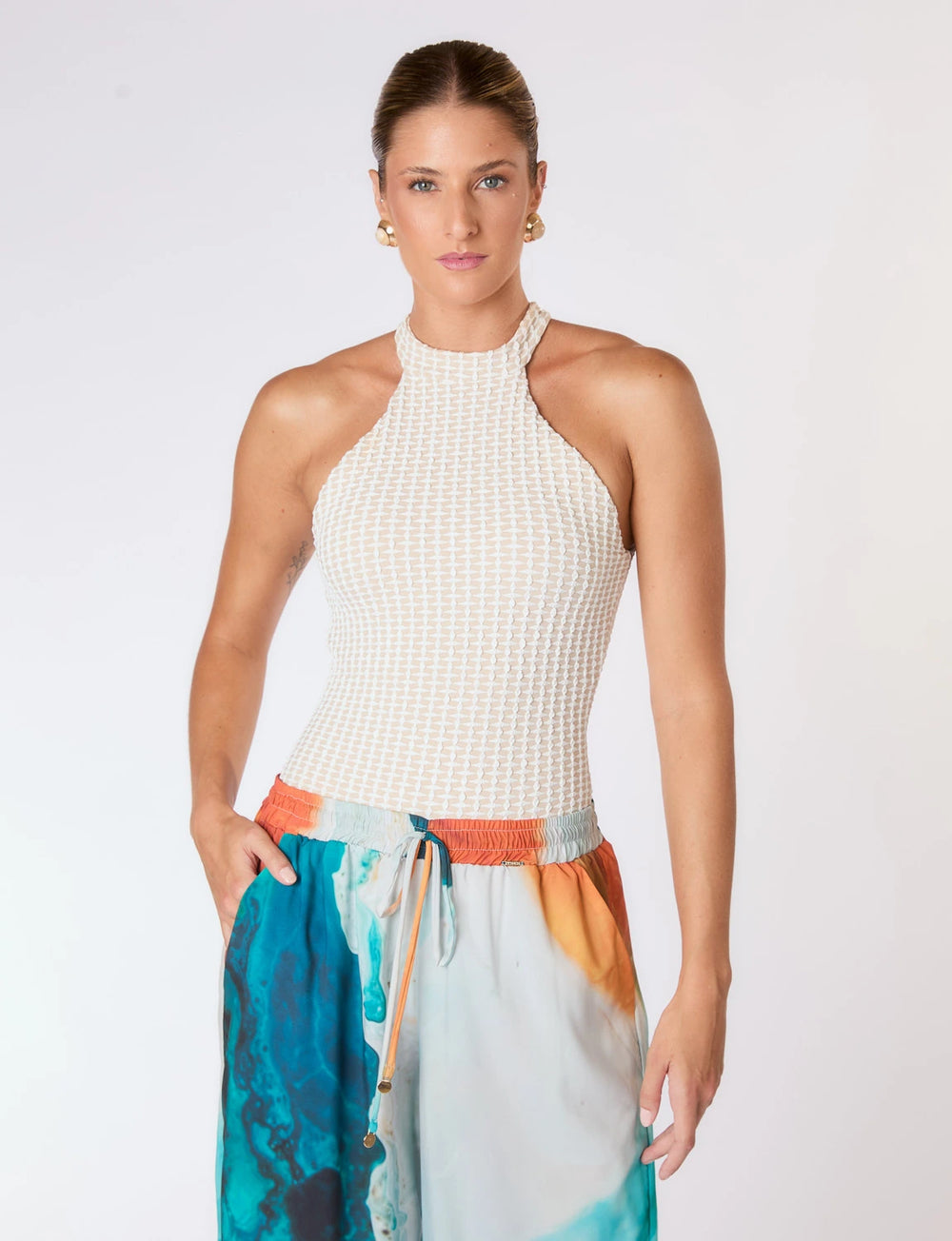 Imagem de mulher usando maiô branco texturizado e calça colorida posando em fundo branco.