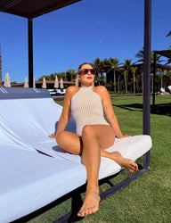 Mulher relaxando em espreguiçadeira ao ar livre, usando óculos de sol, vestindo maiô halter modelador e desfrutando de um dia ensolarado em um resort.