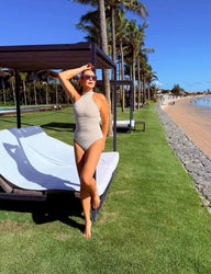 Mulher posando em uma praia tropical usando maiô, com palmeiras ao fundo e céu azul, transmitindo um clima de verão e férias na praia.