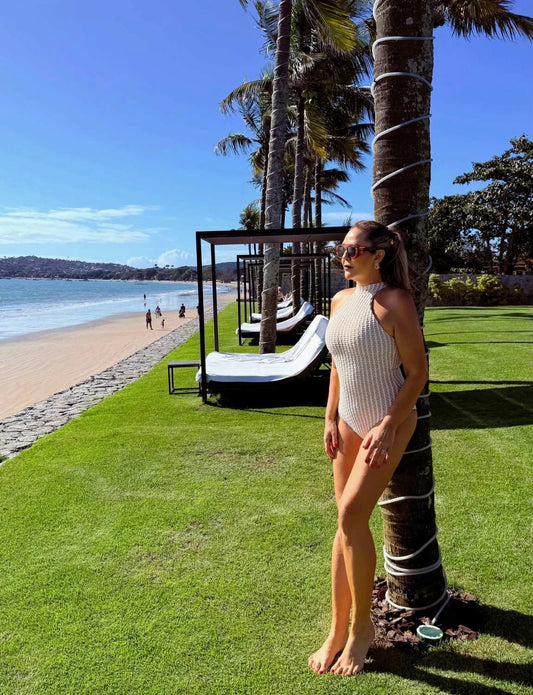 Mulher usando maiô halter textura marroquina com óculos de sol, passeando em praia com espreguiçadeiras de luxo ao ar livre sob palmeiras em tarde ensolarada.