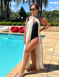 Mulher elegante de cabelo castanho e óculos escuros posando ao lado da piscina em um dia ensolarado, vestindo maiô preto e uma longa peça de roupa branca, com fundo de jardim tropical e mobiliário de lazer.