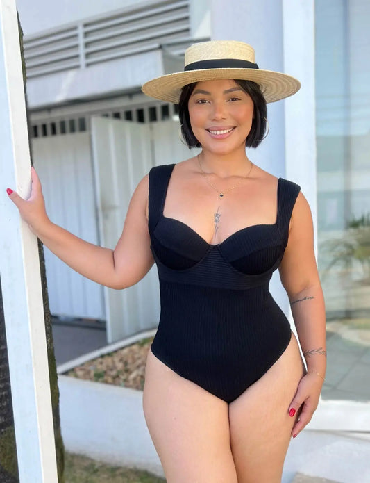 Mulher sorridente com chapéu de palha, usando maiô preto, em ambiente externo, exibindo tatuagens e com cabelo curto e escuro.