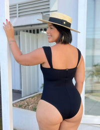 Mulher com cabelo curto, vestindo maiô preto e chapéu de palha, sorrindo enquanto posa ao ar livre. Perfeito para moda praia e estilo verão.