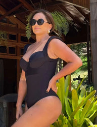 Mulher elegante usando óculos escuros e maiô preto ao ar livre, com plantas e estrutura de madeira ao fundo.