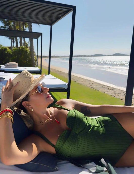 Mulher relaxando na praia deitada ao sol, usando chapéu, óculos de sol e maiô verde, com vista para o mar e céu claro.
