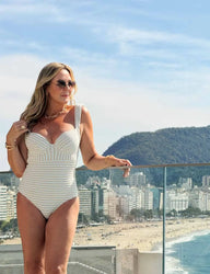 Mulher de cabelos loiros e óculos escuros, usando maiô branco texturizado, em cobertura em frente à praia, com vista para uma montanha ao fundo.