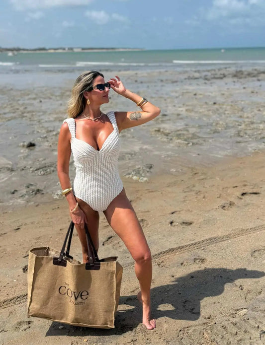 Mulher na praia usando óculos de sol, maiô branco texturizado, carregando uma bolsa com escrito 'Cove'. Fundo com o mar e céu azul com nuvens.