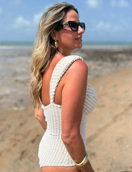 Mulher com cabelos loiros ondulados, usando óculos de sol grandes, brincos dourados e maiô modelador texturizado branco da Kalini Beachwear.