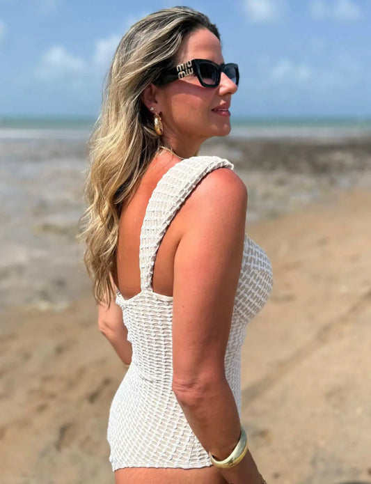 Mulher com cabelos loiros ondulados, usando óculos de sol grandes, brincos dourados e maiô modelador texturizado branco da Kalini Beachwear.