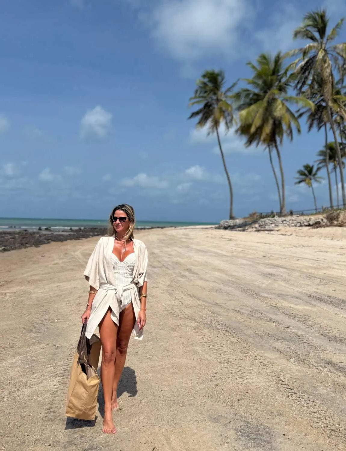 Mulher vestindo maiô modelador branco na praia com coqueiros ao fundo, aproveitando o sol e a areia, em um cenário paradisíaco e tranquilo.