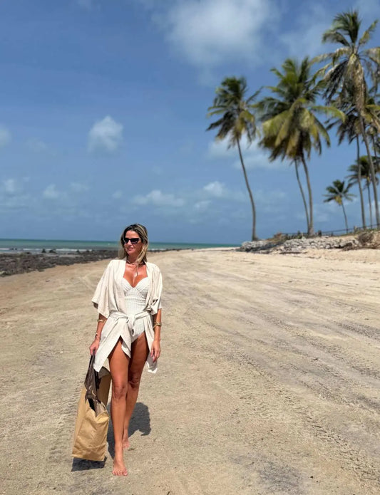 Mulher vestindo maiô modelador branco na praia com coqueiros ao fundo, aproveitando o sol e a areia, em um cenário paradisíaco e tranquilo.