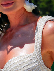 Imagem de mulher ao ar livre com pele queimada pelo sol, usando maiô branco da Kalini Beachwear com detalhes texturizados, destacando o ombro e o pescoço.