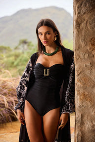 Mulher com maiô preto e acessórios verdes posando ao lado de uma parede de pedra ao ar livre