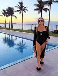 Mulher deslumbrante de óculos escuros e maiô preto com detalhes banhado a ouro, curtindo um pôr do sol e clima de resort à beira com piscina, vista para o mar e palmeiras ao fundo