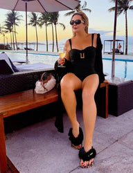 Mulher elegante relaxando à beira da piscina com óculos escuros e maiô preto em ambiente de resort com vista para o mar e palmeiras ao fundo