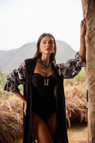 Mulher elegante posando ao ar livre, vestindo body de praia preto com detalhes de tecido transparente e acessórios sofisticados, com fundo natural de montanhas e plantas