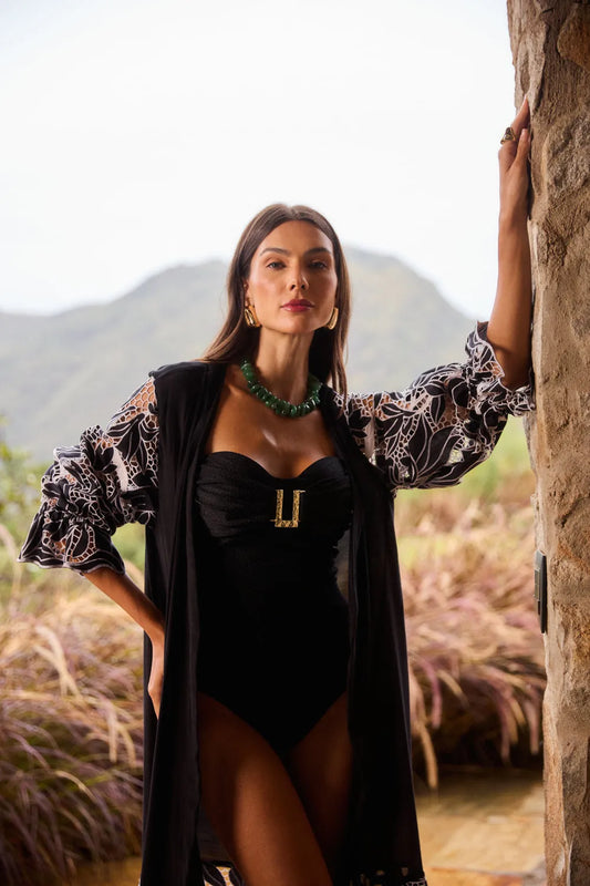 Mulher elegante posando ao ar livre, vestindo body de praia preto com detalhes de tecido transparente e acessórios sofisticados, com fundo natural de montanhas e plantas