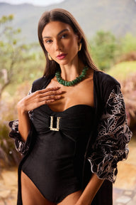 Mulher elegante usando body maiô texturizado preto com detalhes em prata, acessório de colar de pedras verdes e fundo de natureza desfocada